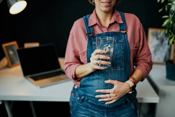 Gravid kvinna som står på sitt hemmakontor, vidrör magen och dricker sötvatten. — Stockfoto