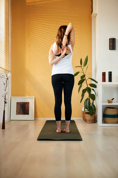Zadní pohled na brunetku středního věku stojící na podložce jógy doma v dopoledních hodinách a natahující paže. — Stock fotografie