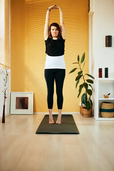 Mid-adult fit slim caucasian brunette standing next to window and stretching arms. Yoga practicing. Morning time. — ストック写真