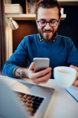 Genç, sakallı hippi evinde oturmuş elinde bir fincan kahveyle akıllı telefon kullanıyor..