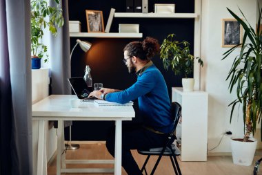 Genç, çekici, beyaz bir hippi ofiste oturuyor ve dizüstü bilgisayarında daktilo yazıyor..