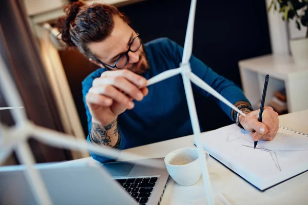 Young creative caucasian bearded environmentalist  with curly hair sitting in his office, touching windmill model and drawing new better model in notebook.