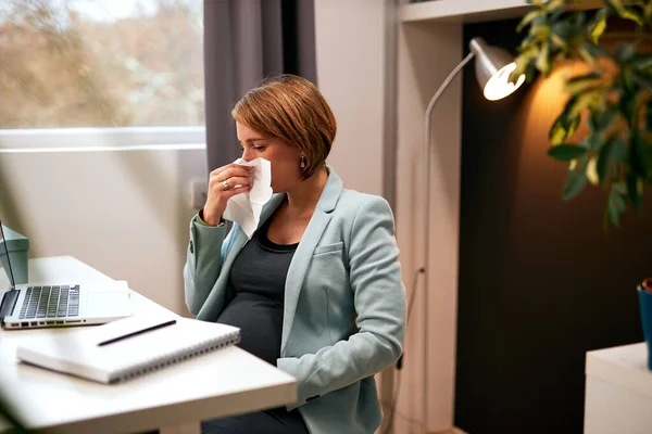 Pregnant businesswoman in forties sitting in her home office sneezing.
