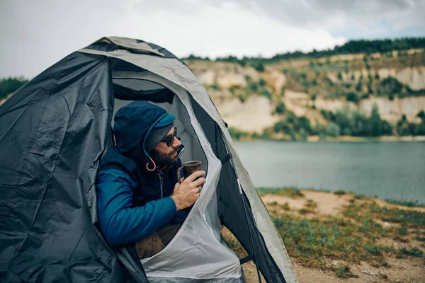 Young handsome bearded man crouching in tent, drinking coffee in the morning and enjoying beautiful nature on camping trip.