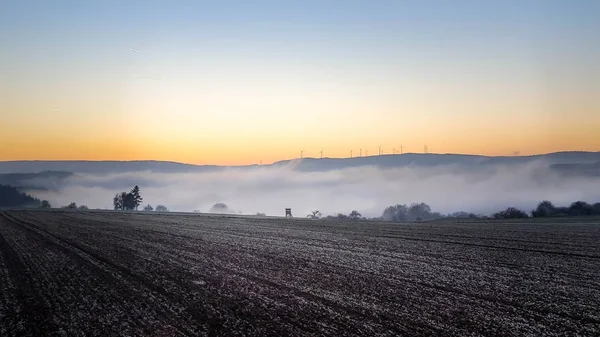 Paysage mélancolique de brouillard tomber dans un champ avec des montagnes et parc éolien pendant le coucher du soleil — Photo