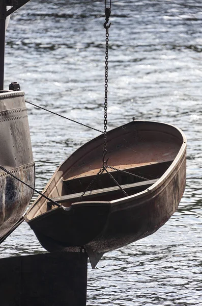 Metallic boat hangs over water on steel chain overboard.
