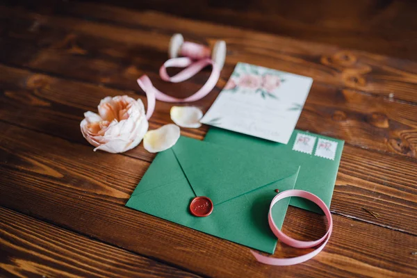 White invitation letter for wedding decorated with flowers and green envelope with seal lying on table with ribbons