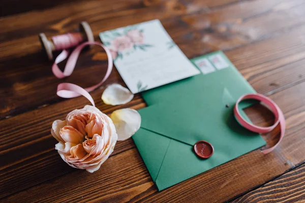 Closeup of white invitation letter for wedding decorated with flowers and green envelope with seal lying on table with ribbons