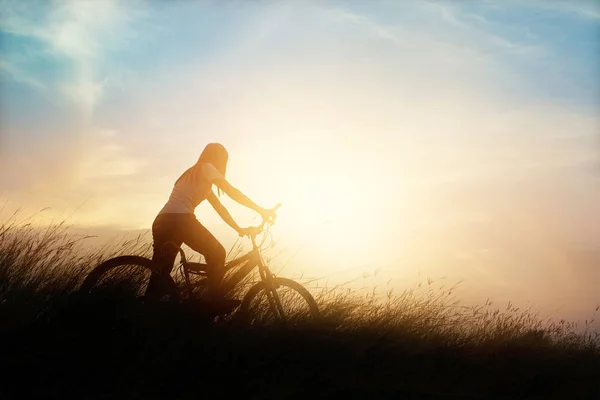 Donna con bicicletta su una strada rurale con erba sullo sfondo del tramonto — Foto Stock