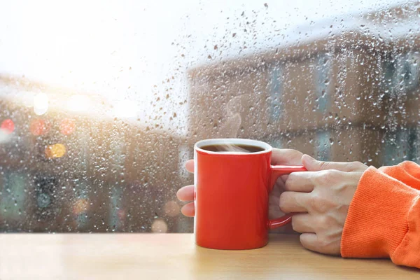 Red coffee cups in hands on a window glass of raindrops background — Stock Photo, Image