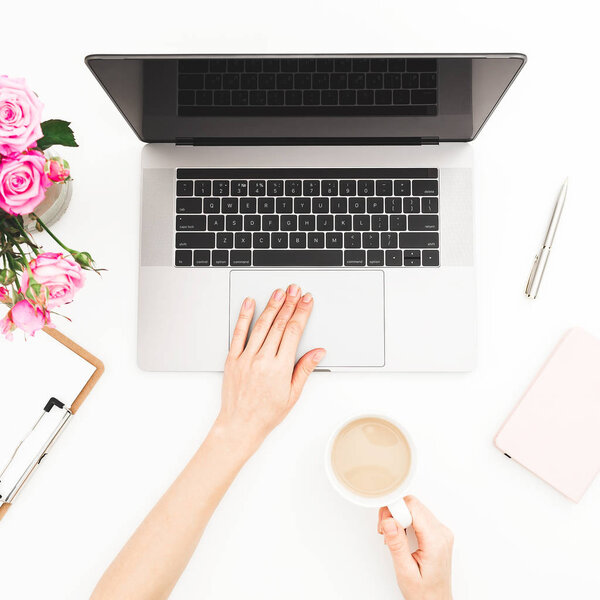 Home office desk. Woman workspace with female hands, laptop, pink roses bouquet, and diary on white background. Flat lay. Top view. Girl working on laptop and hold coffee mug