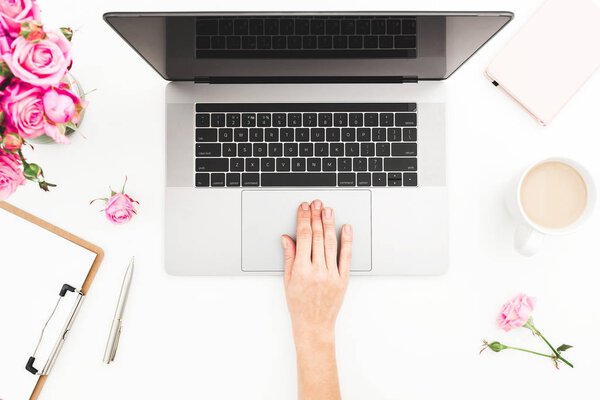 Woman working on laptop. Workspace with female hands, laptop, pink roses bouquet, coffee mug, diary on white table. Top view. Flat lay.