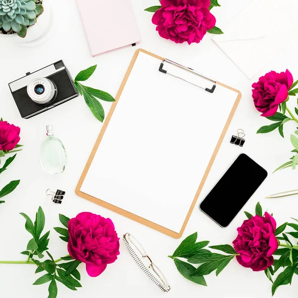 Freelancer workspace with clipboard, dairy, peony flowers, retro camera and smartphone on white background. Flat lay, top view. Beauty blog concept with copy space