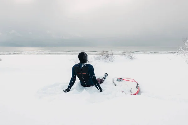 Cold winter and male surfer relax on snow with surfboard. Snowy day with surfer in wetsuit.