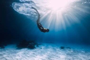Kadın serbest dalgıç kumlu deniz tabanında yüzgeçleriyle süzülüyor. Mavi okyanusta serbest dalış ve güneş ışığı