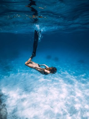 Kadın serbest dalgıç kumlu deniz tabanında yüzgeçleriyle süzülüyor. Mavi okyanusta serbest dalış ve güneş ışığı