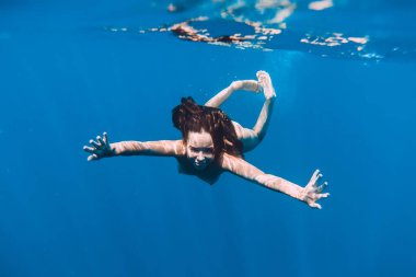 Sıska çıplak kadın şeffaf mavi okyanusta suyun altında poz veriyor. Denizde yüzen çıplak genç kadınlar