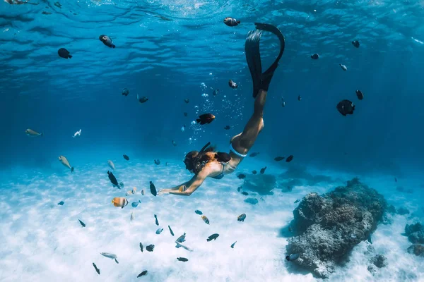 Free diver girl with fins glides over sandy bottom with tropical fishes in blue ocean