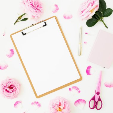 Beyaz zemin üzerinde pembe çiçekler, pano, klipler, defter ve makas olan kompozisyon. Düz yatış, üst görünüm.