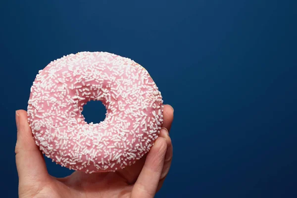 Рука держит вкусный красочный розовый пончик с брызгами рядом с темно-синим фоном, сахарная концепция — стоковое фото