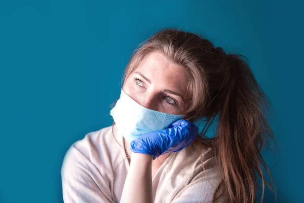 Mujer joven con guantes protectores y mascarilla dentro de una casa en cuarentena que parece aburrida y triste, para Covid-19 Coronavirus, con fondo azul —  Fotos de Stock