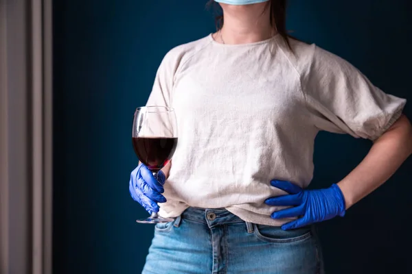 Wanita mengenakan topeng pelindung dan sarung tangan biru di rumahnya dan minum anggur merah, terisolasi untuk Coronavirus Covid-19, sedih dan kesepian — Stok Foto