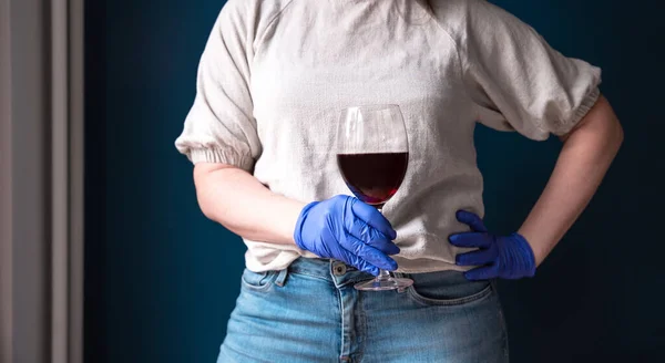 Wanita mengenakan topeng pelindung dan sarung tangan biru di rumahnya dan minum anggur merah, terisolasi untuk Coronavirus Covid-19, sedih dan kesepian — Stok Foto