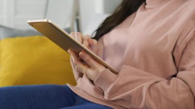 Young woman using rose gold tablet and seat on couch or sofa in jeans and pastel pink sweater. Woman using tablet for social network or shopping online.