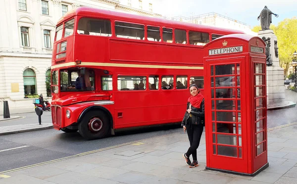 Woman traveling to London, standing near a telephone box and a red british bus.