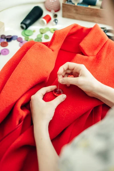 Uma costureira cose um botão em um casaco vermelho na mesa branca — Fotografia de Stock