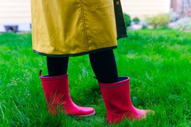 Sarı yağmurluk. Lastik pembe çizmelere karşı. Yeşil çimenlikteki çizmelerle bacakların kavramsal görüntüsü.