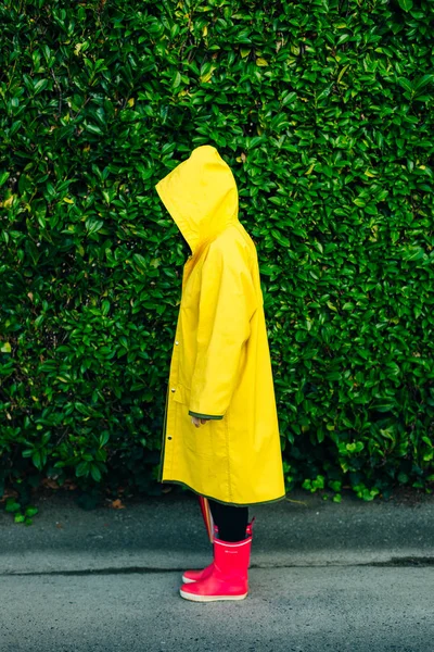 Menina em uma capa de chuva amarela em um fundo de uma parede com grama — Fotografia de Stock