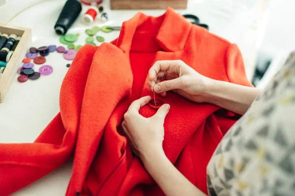 Une couturière coud un bouton sur un manteau rouge sur une table blanche — Photo