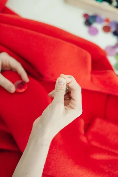 Une couturière coud un bouton sur un manteau rouge sur une table blanche — Photo