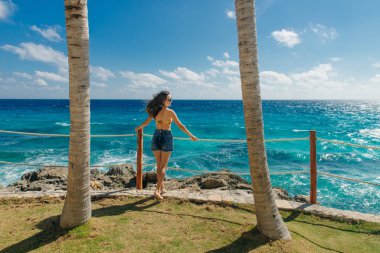 Kız sırtı mavi okyanusa dönük, palmiye ağaçları arasında duruyor. Cancun Meksika