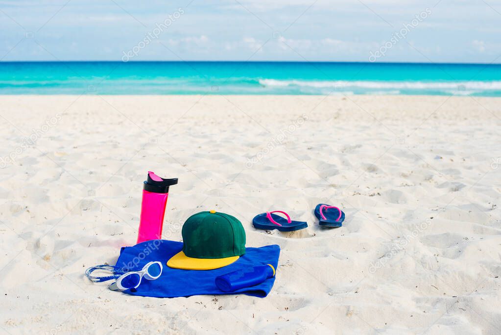 vacation and summer holidays concept - flip flops, towel, cap, bottled water and bottle of sunscreen oil on beach sand