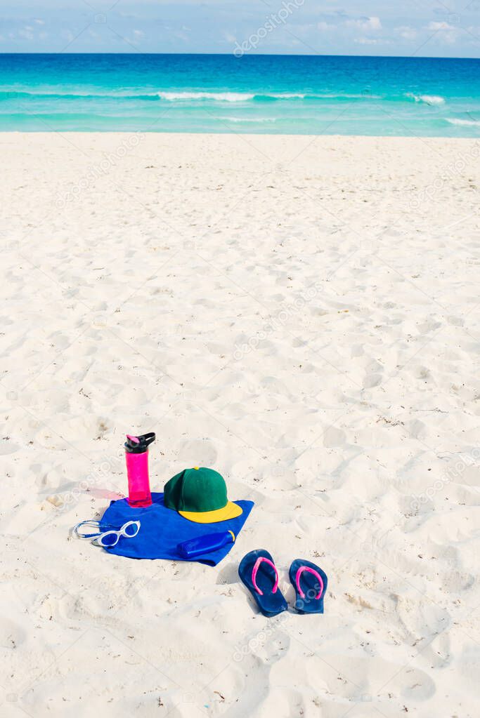 vacation and summer holidays concept - flip flops, towel, cap, bottled water and bottle of sunscreen oil on beach sand