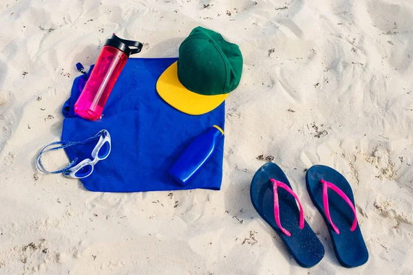 Concepto de vacaciones y vacaciones de verano - chanclas, toalla, gorra, agua embotellada y botella de aceite protector solar en la arena de la playa —  Fotos de Stock