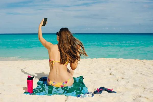 Młoda kobieta w stroju kąpielowym w piasku robi selfie na plaży na swoim smartfonie — Zdjęcie stockowe