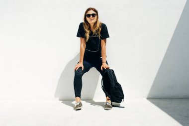 Güneş gözlüklü ve siyah giyen, beyaz arka planda sırt çantalı bir kız..