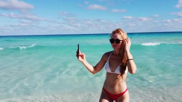 Vista posteriore di una donna in costume da bagno con i capelli lunghi biondi che si fa un selfie su una spiaggia tropicale. 4k — Video Stock