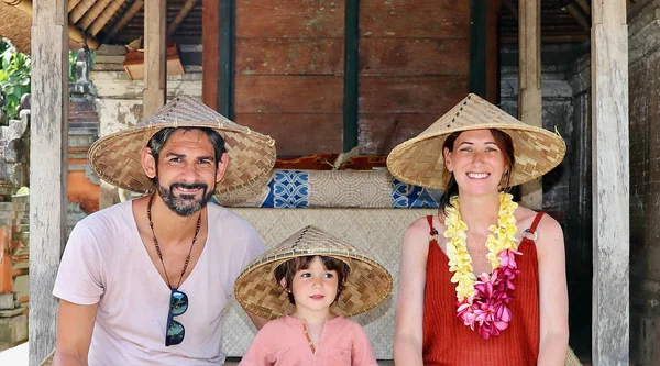 Portrait of a beautiful family of three tourists (two parents and a little kid) wearing the typical balinese hat while visiting a local family home in a village close to Ubud city, Bali, Indonesia.