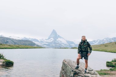 Genç uzun yürüyüşe çıkan kimse bir göl İsviçre Alpleri'nde yanında dinleniyor. Sonraki 