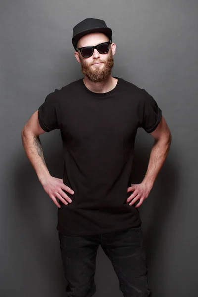 Hipster handsome male model with beard wearing black blank t-shirt with space for your logo or design over gray background — Stock Photo, Image