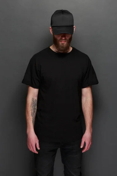 Hipster handsome male model with beard wearing black blank t-shirt and a black baseball cap with space for your logo or design in casual urban style — Stock Photo, Image