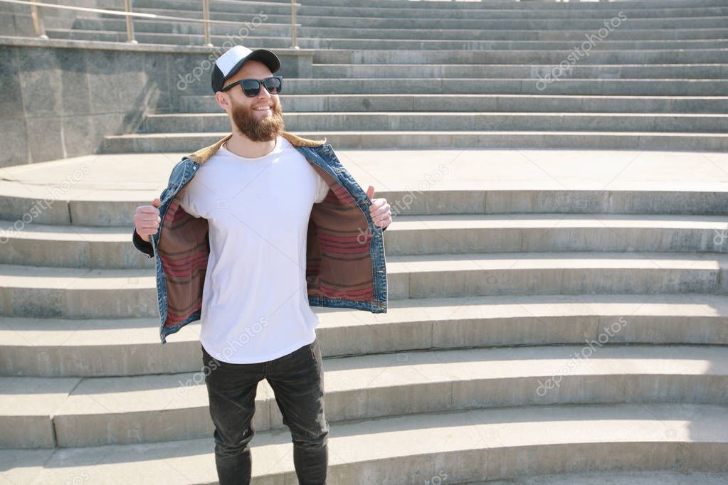 Hipster handsome male model with beard wearing white blank t-shirt and a baseball cap with space for your logo or design in casual urban style