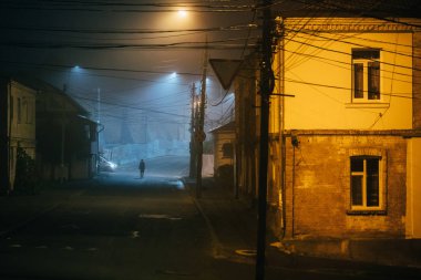 Yalnız bir kadın sisli, eski bir şehirde ceketinde sokak lambalarıyla yürüyor.
