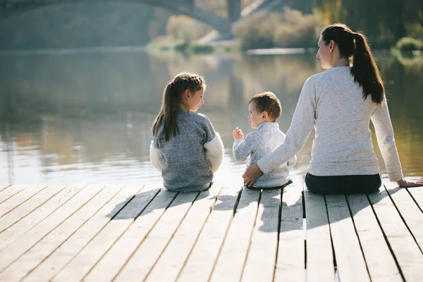 Anya és a gyerekek egy tó mellett pihennek. Mosolyognak és beszélgetnek egy napos őszi napon. — Stock Fotó