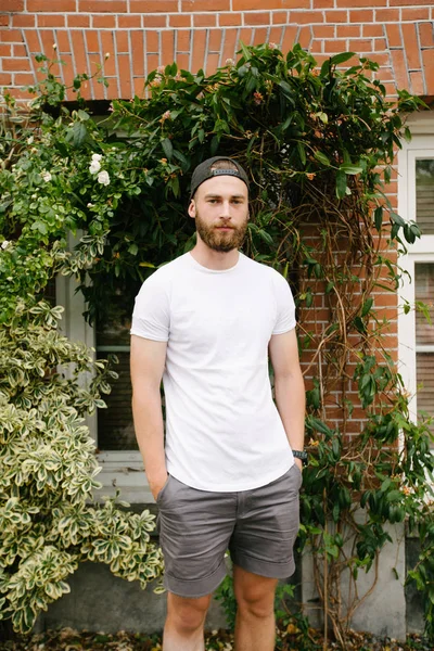 Hipster guapo modelo masculino con barba vistiendo camiseta blanca en blanco y una gorra de béisbol con espacio para su logotipo o diseño en estilo urbano casual — Foto de Stock