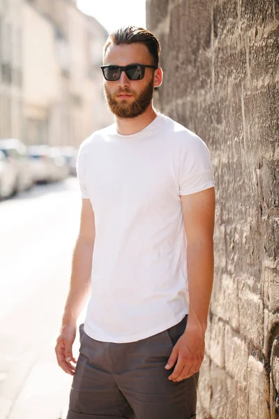 Hipster όμορφο αρσενικό μοντέλο με γενειάδα φορώντας λευκό κενό t-shirt και ένα καπέλο του μπέιζμπολ με χώρο για το λογότυπο ή το σχέδιό σας σε casual αστικό στυλ — Φωτογραφία Αρχείου
